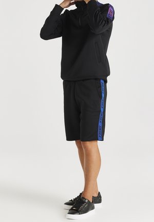 Sort lynlås-sweatshirt med høj krave, kombineret med sorte shorts med en blå stribe i siden. Sorte sneakers med gyldne detaljer.