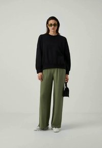 Felpa nera abbinata a pantaloni larghi verde oliva. Il modello indossa occhiali da sole verdi e tiene una piccola borsa nera. Scarpe da ginnastica bianche.