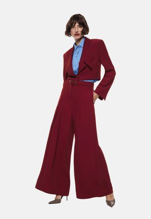 Donna in posa con pantaloni a gamba larga color bordeaux, blazer corto, camicia a righe blu e scarpe con punta metallica su uno sfondo bianco.