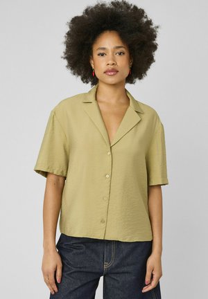 Femme avec une coiffure afro naturelle portant une chemise à manches courtes couleur olive clair et un jean bleu foncé, debout devant un fond uni.
