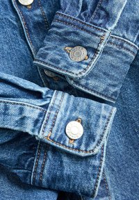 Blue denim shirt cuffs featuring silver snap buttons, contrasting orange stitching, and slightly frayed edges with a textured finish.
