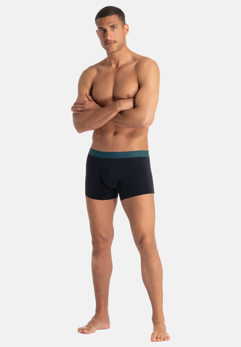 Czarne bokserki z turkusowym paskiem w talii, cechujące się przylegającym krojem i miękką fakturą, prezentowane przez osobę stojącą z założonymi na siebie ramionami.