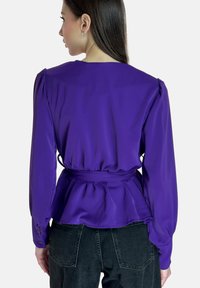 Blouse peplum violette avec manches longues, taille ajustée et poignets boutonnés, présentée sur fond gris clair.