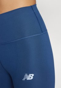Tamno plave atletske tajice izrađene od rastezljivog materijala s glatkom teksturom. Sadrže ušivenu traku oko struka i bijeli New Balance logotip.