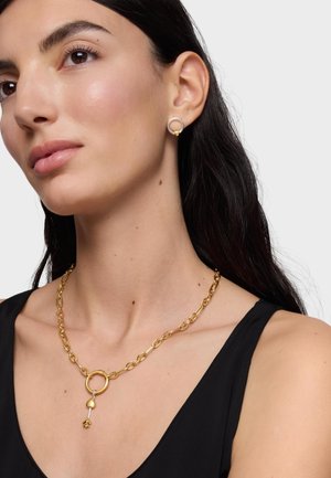 Kobieta z długimi ciemnymi włosami, nosząca złoty łańcuszek z zawieszką w kształcie serca i strzały oraz srebrne kolczyki w kształcie okręgów, ubrana w czarną bluzkę.