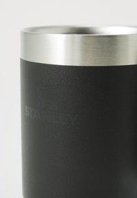 Tasse en acier inoxydable noir avec un bord en métal brossé, dotée d'une surface texturée et du logo "STANLEY" en relief.
