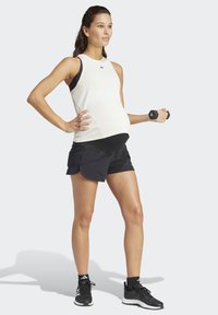 Top de tirantes blanco con detalles negros, pantalones cortos deportivos negros, sosteniendo una pesa, con una postura recta y una ligera sombra sobre un fondo neutral.
