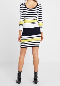 Robe en maille côtelée avec rayures horizontales bleu marine, blanc et jaune. Conçue avec des manches longues et un dos dégagé. Bottines noires.