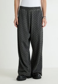 Black checkered sweatpants with an elastic waistband, loose fit, and subtle texture; paired with black studded slip-on shoes.