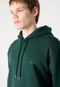 Sudadera verde con capucha y acabado texturizado, que presenta cordones y un logo impreso en un verde más claro en el pecho.