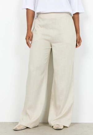 Frau trägt hochtaillierte, weit geschnittene beige Hose, weißen Oberteil und flache gemusterte Schuhe, steht vor einer weißen Wand.