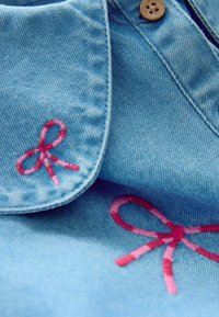 Denimstof med lyseblå tekstur, prydet med lyserøde broderede sløjfer på kraven og kroppen, samt en detalje med en træknap.