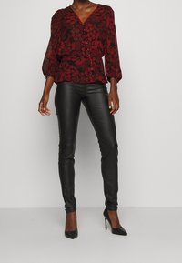 Blusa floreale rossa e nera con scollatura a V, dettagli con bottoni e maniche a sbuffo, abbinata a pantaloni di pelle nera e scarpe nere con tacco alto.