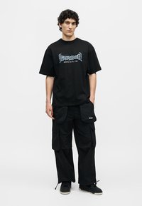Jeune homme debout portant un t-shirt graphique noir avec le texte « Barrow Greatest of All Time », un pantalon cargo noir et des baskets noires sur fond blanc.