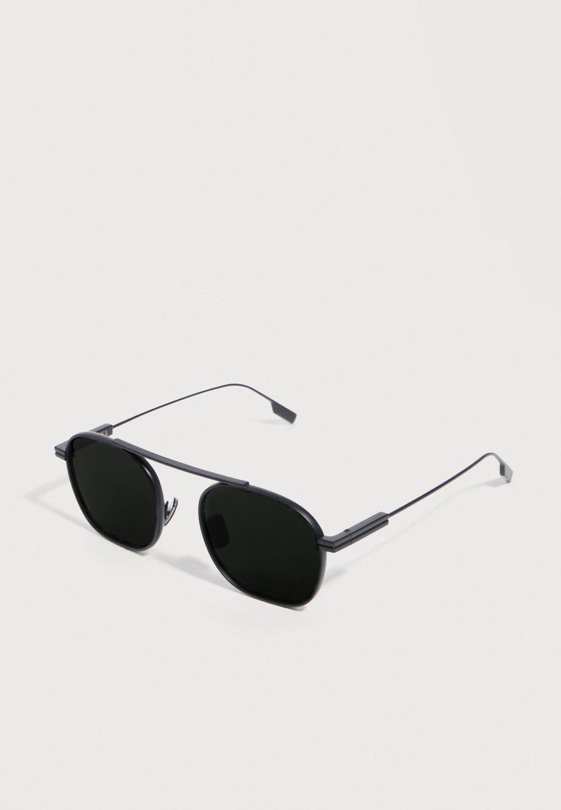 Black metal-framed sunglasses with thin arms and dark tinted lenses, resting on a white surface.