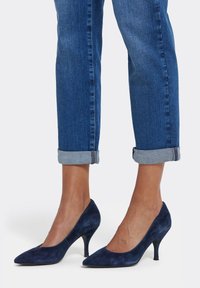 Mörkblå mockaskor med hög klack, spetsig tå och en stilettklack på 4 cm, kombinerade med uppvikta jeans. Slät textur, minimalistisk design.