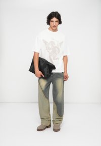Balta per didelį t-shirt su sparnuotu cherubu spaudu, nusidėvėjusi dviejų tonų džinsai su išpruvusiais kraštais ir juoda rankinė; modelis vilki beželes batus.