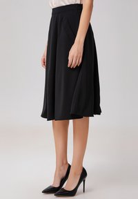 Jupe noire en A fabriquée en tissu lisse, avec des poches latérales et une fermeture éclair dissimulée. Associée à des escarpins noirs à bout pointu.