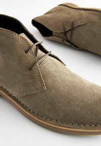 Botas de ante hasta el tobillo en verde oliva con una textura suave, que presentan punteras redondas, costuras marrones en contraste y cordones marrones.