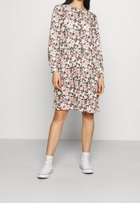 Robe florale à manches longues, principalement noire avec des fleurs roses et vertes. Coupe ample, longueur genoux, associée à des baskets montantes blanches.