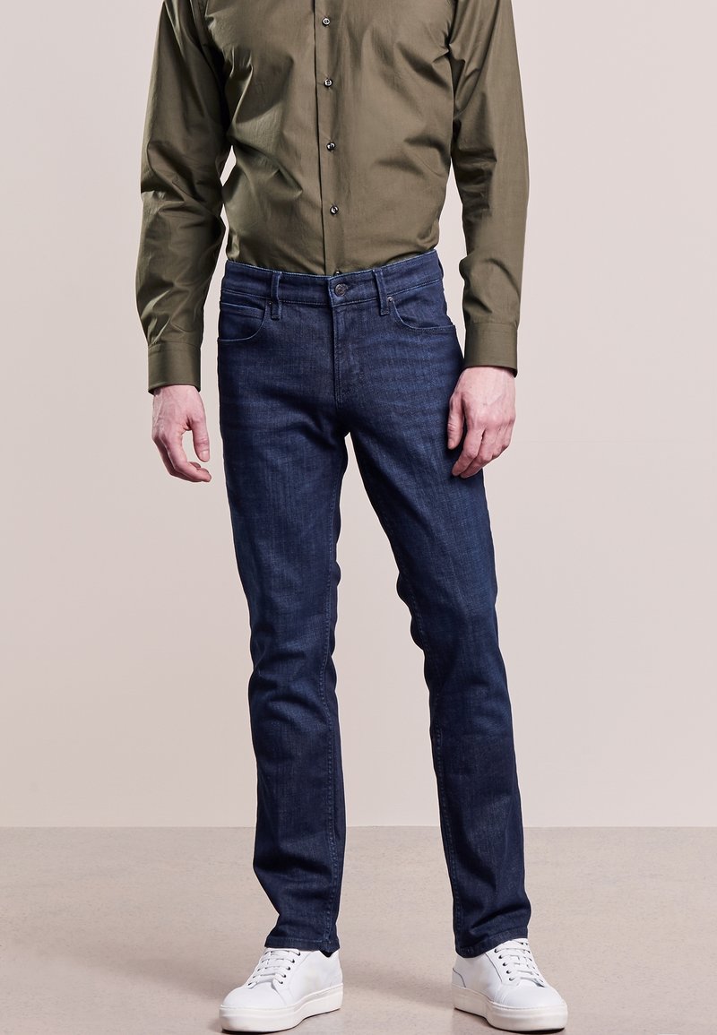 Man som bär mörkblå slim-fit jeans, olivgrön knäppt skjorta och vita sneakers står på en ljus beige golv.