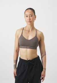 Barna sportmelltartó V-alakú dekoltárral és vékony pántokkal. A megfelelő árnyalatú Nike logóval ellátott pántja van. A modellen kényelmes fekete melegítőnadrág van.