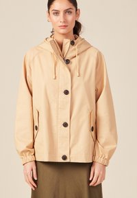 Giacca cappuccio di un leggero beige con chiusura a bottoni e polsini elastici, dotata di due tasche laterali e un design semplice. Texture liscia.