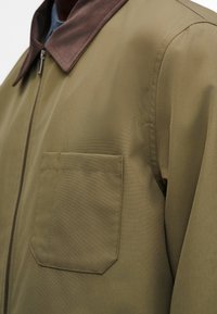 Gros plan sur une veste kaki avec un col marron foncé, une fermeture éclair visible à l'avant et une poche poitrine sur le côté gauche.