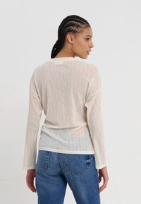 Lange mouwen, lichtgewicht top in beige met een doorschijnende textuur en geribbeld patroon. De achterkant heeft een lichte drop en een relaxte fit. Gecombineerd met blauwe jeans.
