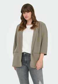 Blazer khaki de corte abierto con mangas de tres cuartos, que cuenta con dos bolsillos frontales y un ajuste ligeramente holgado, llevado sobre una camiseta blanca y unos jeans gris.