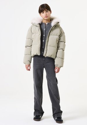 Doudoune kaki avec capuche en fausse fourrure, design matelassé, poches zippées, portée sur un pull gris et associée à un jean gris et des chaussures noires.