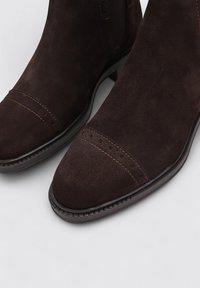 Bottines en suede marron avec une texture lisse, un bout arrondi, des surpiqûres contrastantes et des perforations décoratives sur la partie avant.