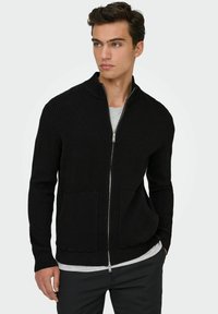 Crna rebrasta zip-up kardigan s visokim ovratnikom, koji ima dva prednja džepa i teksturiranu tkaninu. Nosi se preko sive majice.