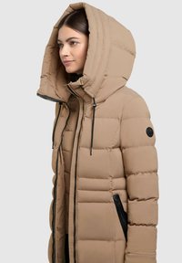 Beige párnázott dzseki nagy kapucnival, elöl cipzárral, húzózsinórral és fekete cipzáras oldalzsebekkel. Az anyag textúrázott steppelt mintázattal rendelkezik.