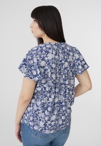 Blusa floreale blu con maniche corte, caratterizzata da uno scollo arricciato sul retro e motivi floreali bianchi su un tessuto leggero. Abbinata a jeans in denim.