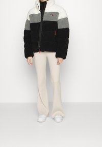 Giacca in pile con strisce orizzontali bianche, grigie e nere, dotata di zip e accenti rossi. Abbinata a pantaloni a coste beige chiaro.
