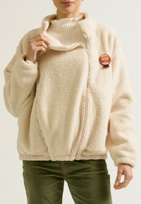 Cremefarbene Fleecejacke mit hohem Kragen, Reißverschluss und einem "Mama"-Abzeichen. Der Stoff hat ein weiches, strukturiertes Aussehen.