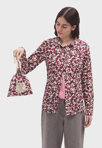Camisa de patrón floral colorida en rosa, rojo y blanco, con mangas largas y cierre de botones en el frente. El modelo sostiene una pequeña bolsa de cordón.