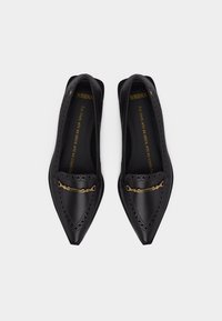 Mocassins en cuir noirs à bouts pointus avec perforations décoratives et détail en métal doré en forme de mors de cheval sur le dessus.