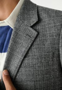 Grijze geblokte blazer met een gestructureerd ruitpatroon. De stof heeft een subtiele glans. Een wit overhemd met blauwe strepen is zichtbaar eronder.