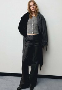 Cardigan scurt gri cu nasturi, combinat cu pantaloni negri evazați. O eșarfă lungă neagră cu franjuri se așază pe o umăr. Texturi netede.