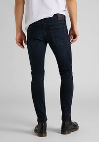 Vaqueros de corte ajustado de mezclilla azul oscuro con cinco bolsillos, trabillas para cinturón y un parche de cuero negro texturizado en la parte trasera de la cinturilla.