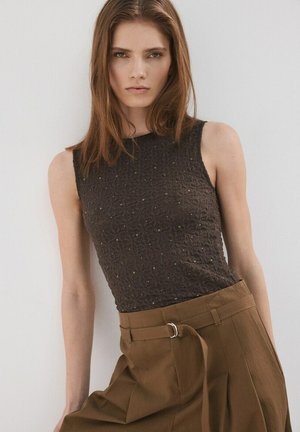 Femme aux cheveux bruns lisses vêtue d'un haut noir sans manches texturé et d'un pantalon marron taille haute avec une ceinture, debout contre un mur blanc.