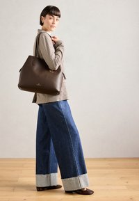 Sac tote en cuir marron avec un design structuré, présentant une texture lisse et des côtés effilés. Porté avec un jean en denim à large jambe et un blazer à carreaux.
