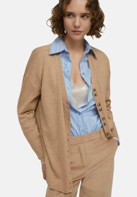 Cardigan beige dal taglio morbido, con bottoni lungo la parte anteriore. Indossato sopra una camicia a righe blu e pantaloni in corduroy beige.
