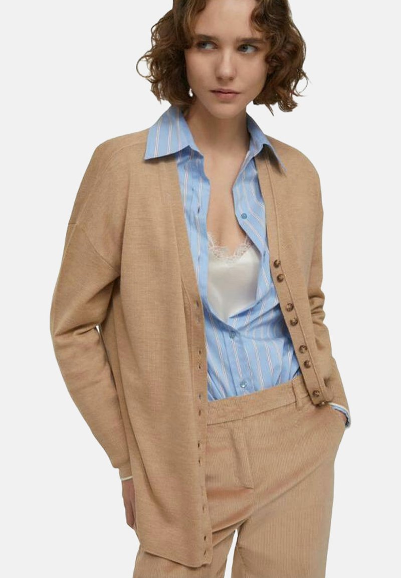 Cardigan beige dal taglio morbido, con bottoni lungo la parte anteriore. Indossato sopra una camicia a righe blu e pantaloni in corduroy beige.