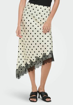 Jupe midi crème à pois noirs avec bordure en dentelle, ourlet asymétrique, portée avec des sandales à talons noires et un haut noir.