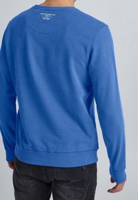 Sudadera de algodón azul con mangas largas, puños y dobladillo acanalados, y un cuello redondo. Presenta un detalle de texto en la parte trasera cerca del cuello.
