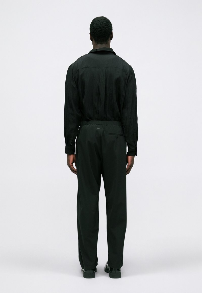 Mand står med ryggen til, iført sort langærmet skjorte, sorte bukser med elastisk linning og sorte sko på en ensfarvet lys baggrund.