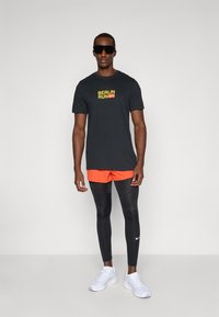 Black t-shirt with "BERLIN RUN" in yellow and red, orange shorts, black leggings, and white athletic shoes. Square sunglasses complete the look.